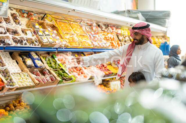 تعليم الأبناء أنواع الفاكهه والخضروات المعروضه على أرفف الثلاجه في الماركت، إستشارة الإبنة في شراء السلع والمستلزمات المنزلية، أب عربي خليجي سعودي يرتدي الشماغ والثوب التقليدي ويقضي وقت ممتع برفقة إبنته في إختيار أفضل الفواكه الطازجة من السوبرماركت.