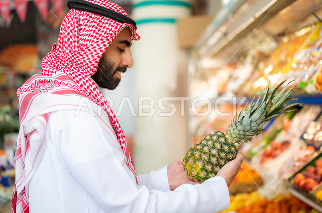 Selecting the best types of fruits from the supermarket, gestures of pleasure and happiness when shopping and buying consumer supplies from the food store, a close-up picture of a Saudi Gulf Arab man wearing a shemagh and traditional thobe holding a pineapple in the fruits and vegetables section, Saudi agricultural crops, national local products.