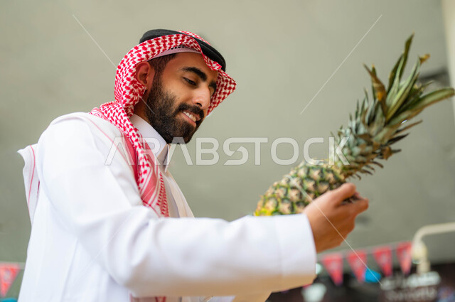 انتقاء أفضل أنواع الفاكهة من السوبرماركت، إيماءات السرور والسعادة بالتسوق وشراء مستلزمات المستهلك من متجر المواد الغذائية، صوره مقربه لرجل عربي خليجي سعودي يرتدي الشماغ والثوب التقليدي يحمل حبة الأناناس في قسم الفواكه والخضار، محاصيل زراعية سعودية, منتجات محلية وطنية.