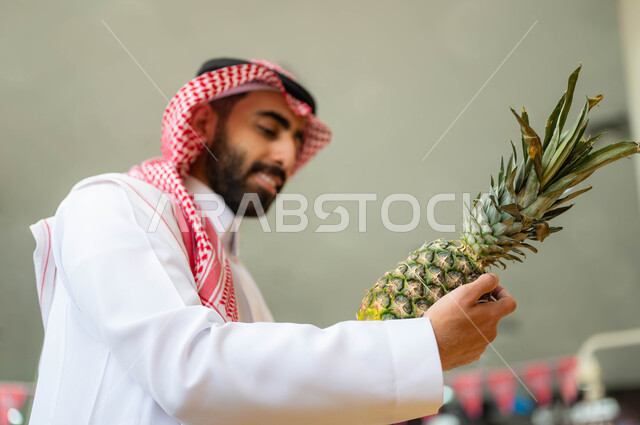 إيماءات السرور والسعادة بالتسوق وشراء مستلزمات المستهلك من متجر المواد الغذائية، صوره مقربه لرجل عربي خليجي سعودي يرتدي الشماغ والثوب التقليدي يحمل حبة الأناناس في قسم الفواكه والخضار، انتقاء أفضل أنواع الفاكهة من السوبرماركت، محاصيل زراعية سعودية, منتجات محلية وطنية.