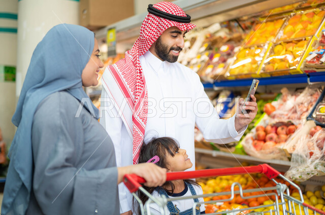 مشاركة الأسرة في توفير وشراء اللوازم والإحتياجات المنزلية، الإعجاب بجودة الفواكه والخضراوات، منتجات محلية وطنية، رجل عربي خليجي سعودي برفقة عائلته يقوم بتصوير الفاكهة الطازجة في السوبرماركت بإستخدام الهاتف المحمول، محاصيل زراعية سعودية.