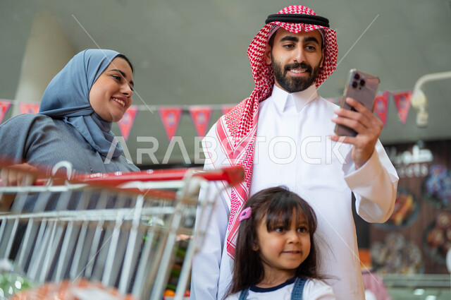 مفهوم التسوق العائلي وشراء الإحتياجات والمستلزمات المنزلية، رجل عربي خليجي سعودي برفقة عائلته يقوم بإجراء مكالمة فيديو في السوبرماركت من خلال الهاتف المحمول، استخدام الجوال في مشاهدة عروض وخصومات الجمعة البيضاء، إيماءات السعادة والسرور أثناء التسوق.