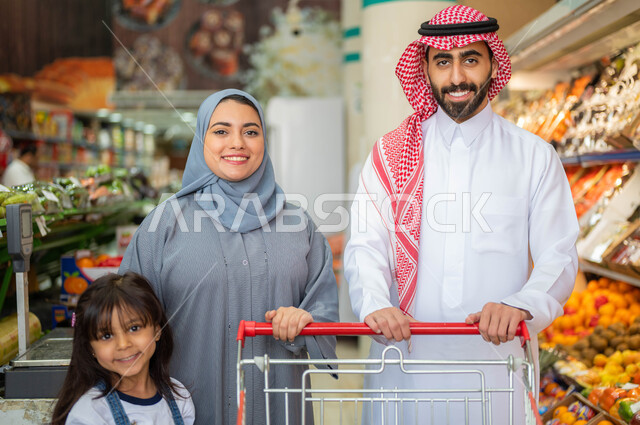 ايماءات الفرح والسعاده لقضاء أوقات ممتعه برفقه العائله، الاستعداد للتسوق وشراء المستلزمات من إحدى المتاجر الغذائية، عائلة عربيه خليجيه سعوديه تبتسم وتقوم بالنظر إلى الكاميرا في قسم الخضراوات بالسوبرماركت، الاستفاده من التخفيضات والعروض على المنتجات.