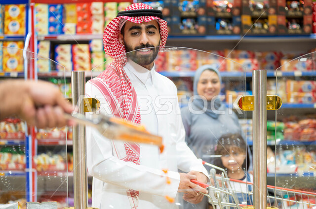 التعاون ومشاركة العائلة في إختيار أنواع مختلفة من المكسرات من الماركت، قضاء الوقت في التسوق برفقة العائلة، أسرة عربية خليجية سعودية تقوم بشراء التسالي والمقرمشات الطازجه من السوبرماركت، استغلال العروض في توفير الاحتياجات المنزلية.