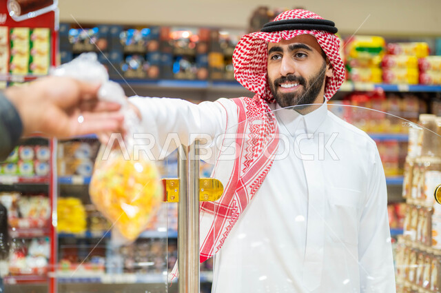 شراء الأحتياجات المنزليه من قسم المكسرات في السوبرماركت، مستلزمات المستهلك من سلع ومواد غذائيه مختلفه في التجر الغذائي، رجل عربي خليجي سعودي مبتسم ويرتدي الزي السعودي التقليدي يقوم بأخذ كيس المقرمشات والتسالي من البائع، وجبات وسناكات صحيه خفيفه.