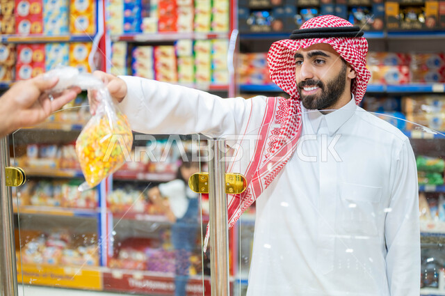 Consumer supplies of various goods and foodstuffs in food stores. A smiling Saudi Gulf Arab man wearing traditional Saudi clothing takes a bag of crackers and snacks from the seller, purchasing household needs from the nuts section of the supermarket, and light healthy meals and snacks.