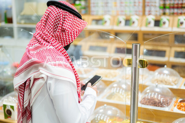 Ensuring the purchase of the required goods on the list of supplies, a picture from behind of a Saudi Gulf Arab man wearing a shemagh and traditional thobe, checking six purchases over the phone, shopping from the nuts section in the food store, taking advantage of the offers and discounts in the Saudi supermarket.