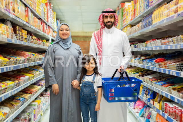 ايماءات الفرح والسعادة بالتسوق وشراء المستلزمات، عائلة عربية خليجية سعودية مبتمسون ينظرون إلى الكاميرا، رجل سعودي يرتدي الثوب والشماغ يمسك بيده سلة التسوق، إمراة سعوديه مبتمسة ترتدي العباءة الرمادية والحجاب تمسك بيد ابنتها، قضاء وقت ممتع في السوبرماركت.