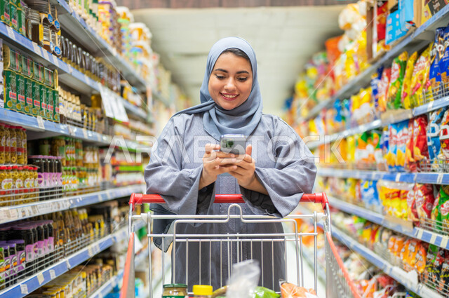 أرفف المواد الغذائيه والسلع المتنوعة في أقسام الماركت، الإستمتاع بشراء الاحتياجات واللوازم المنزلية من السوبرماركت، امرأة عربيه خليجية سعودية محجبة تقوم بجر عربه التسوق وقراءه قائمة الطلبات بإستخدام الهاتف المحمول، الاستفادة من العروض والخصومات على المنتجات السعوديه.