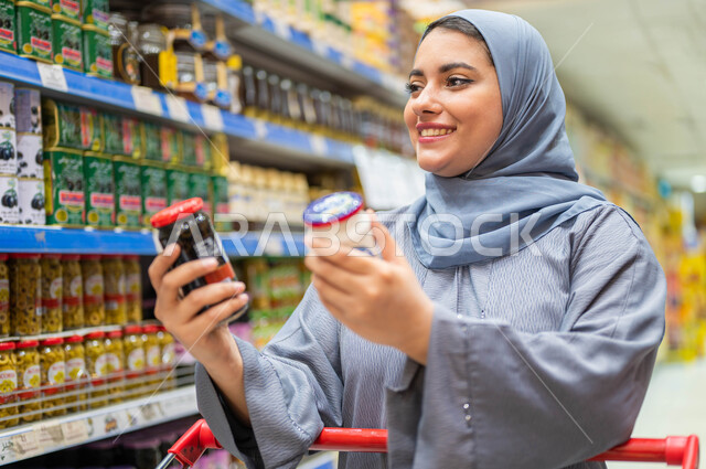 الاستفاده من تخفيضات الجمعه البيضاء، انتقاء واختيار أفضل وأجود السلع والمنتجات من السوبرماركت، شابه عربيه خليجيه سعوديه محجبه ترتدي عباءه رماديه وتقوم بالمقارنه بين نوعين من المنتجات في يدها.