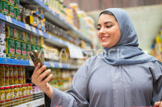 الاستفادة من العروض على المنتجات السعوديه، التأثير الإيجابي للتسوق على النساء, صوره مقربه لامرأه عربيه خليجية سعودية محجبة تقوم بقراءة قائمة المشتريات من الجوال بإيماءات السعادة والسرور، شراء السلع والمواد الغذائيه المتنوعة في الأقسام.