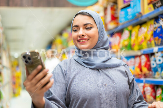 التأثير الإيجابي للتسوق على النساء, صوره مقربه لامرأه عربيه خليجية سعودية محجبة تقوم بقراءة قائمة المشتريات من الجوال بإيماءات السعادة والسرور، شراء السلع والمواد الغذائيه المتنوعة في الأقسام، الاستفادة من العروض على المنتجات السعوديه.