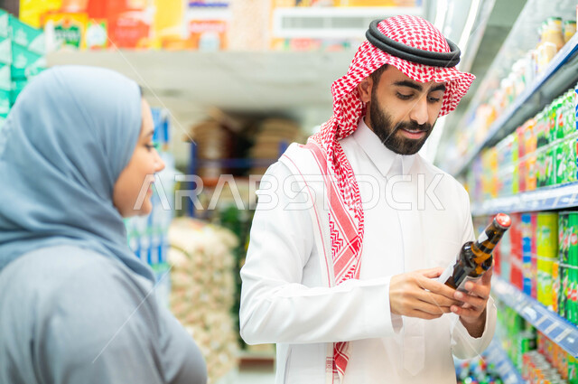 الاستفادة من التخفيضات والعروض والخصومات على المنتجات، زوجان عربيان خليجيان سعوديان مبتسمان يقومان بالتأكد من تاريخ صلاحية المنتج ومحتوياته، سيدة خليجية ترتدي العباءة الرمادية، رجل سعودي يرتدي الثوب والشماغ، الاستعداد للتسوق وشراء المستلزمات.