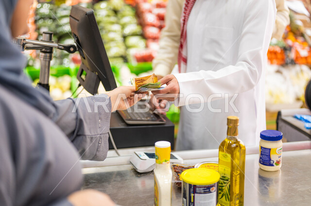 متعة التبضع وشراء السلع المختلفة من متجر المواد الغذائيه، شراء الاحتياجات المنزليه ومستلزمات المستهلك من السوبرماركت، صوره مقربة لإمرأة عربيه خليجيه سعودية تقوم بالدفع النقدي للمحاسب على فاتوره المشتريات، منتجات محليه وطنية، الإستفادة من العروض والتخفيضات.