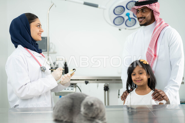 طبيبة بيطرية مبتسمة ترتدي الحجاب والمريول الابيض تقوم بمعالجة وفحص قطة رمادية صغيرة، عائلة عربية خليجية سعودية تقوم بزيارة المراكز والعيادات البيطرية السعودية، أهمية الفحص الطبي الدوري للقطط المنزلية, مفهوم الاهتمام والعناية بالحيوانات الاليفة