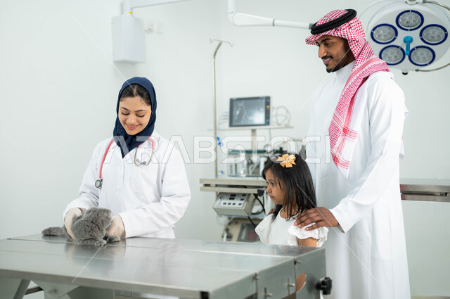 طبيبة بيطرية مبتسمة ترتدي الحجاب والمريول الابيض تقوم بمعالجة وفحص قطة رمادية صغيرة، عائلة عربية خليجية سعودية تقوم بزيارة المراكز والعيادات البيطرية السعودية، أهمية الفحص الطبي الدوري للقطط المنزلية, مفهوم الاهتمام والعناية بالحيوانات الاليفة