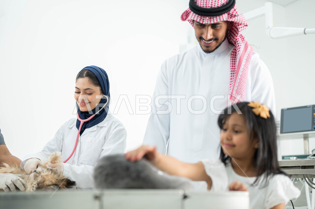 مفهوم الاهتمام والعناية بالحيوانات المنزلية، ضرورة الفحص الطبي الدوري للحيوانات الأليفة, طبيبة بيطرية محجبة تقوم بفحص ومعالجة كلب صغير، رجل سعودي عربي خليجي يرتدي الثوب التقليدي والشماغ يقوم بزيارة المراكز والعيادات البيطرية برفقة ابنته