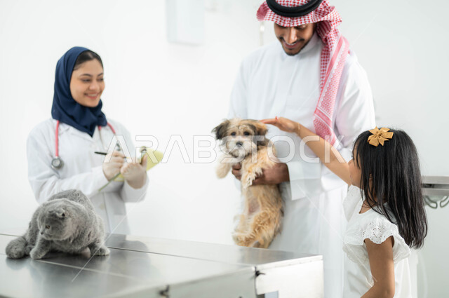 تعامل الأطفال بلين ولطف مع القطط والكلاب, مفهوم الرفق والاعتناء بالحيوان , أب سعودي عربي خليجي مبتسم يرتدي الثوب التقليدي يقوم بزيارة العيادة البيطرية برفقة ابنته,  طبيبة بيطرية محجبة تقوم بتدوين معلومات وبيانات الحيوانات