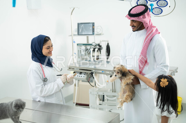 تعامل الأطفال بلين ولطف مع القطط والكلاب, مفهوم الرفق والاعتناء بالحيوان , أب سعودي عربي خليجي مبتسم يرتدي الثوب التقليدي يقوم بزيارة العيادة البيطرية برفقة ابنته,  طبيبة بيطرية محجبة تقوم بتدوين معلومات وبيانات الحيوانات