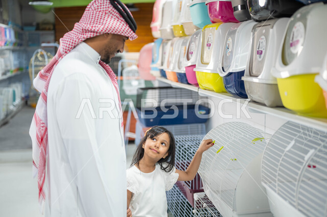 اختيار صندوق نقل الحيوانات الأليفة من متجر بيع وشراء الحاجات والمستلزمات الخاصة بالحيوان، قضاء اوقات ممتعة ومسلية في التسوق مع الأبناء، فتاة عربية خليجية سعودية تشير إلى قفص الهامستر المنزلي بجانب والدها