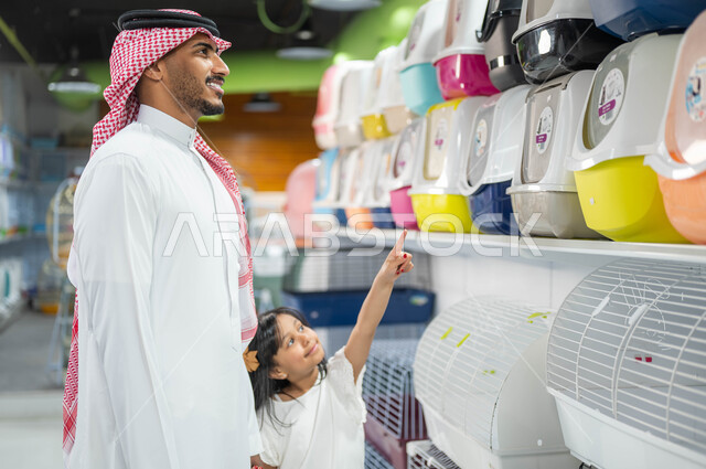 اختيار صندوق نقل الحيوانات الأليفة من متجر بيع وشراء الحاجات والمستلزمات الخاصة بالحيوان، فتاة عربية خليجية سعودية تشير إلى قفص الهامستر المنزلي بجانب والدها، قضاء اوقات ممتعة ومسلية في التسوق مع الأبناء