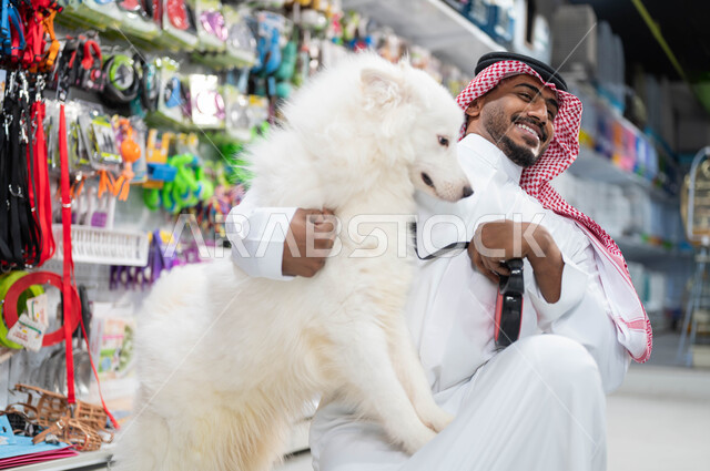 ايماءات السعادة والسرور لقضاء وقت ممتع في اللعب مع الحيوان الأليف، حب الكلاب للنشاط والحركة الدائمة، صورة مقربة لرجل عربي خليجي سعودي مبتسم يرتدي الزي السعودي التقليدي يجلس جانب كلبه الأبيض في متجر المستلزمات والحاجات الخاصة بالحيوانات الأليفة 