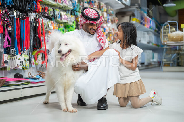 مفهوم الرفق بالحيوان الودود المخلص، رجل عربي خليجي سعودي مبتسم يرتدي الزي السعودي التقليدي يجلس برفقة ابنته الصغيرة، متاجر بيع وشراء حاجات مستلزمات الحيوانات الأليفة، ايماءات السعادة والمرح واللعب مع الكلب الأبيض الوفي