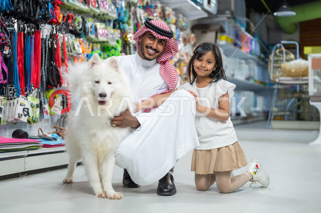 متاجر بيع وشراء حاجات ومستلزمات الحيوانات الأليفة، رجل عربي خليجي سعودي مبتسم يرتدي الزي السعودي التقليدي ينظر إلى الكاميرا يجلس برفقة ابنته الصغيرة، ايماءات السعادة والمرح واللعب مع الكلب الأبيض الوفي، مفهوم الرفق بالحيوان الودود المخلص