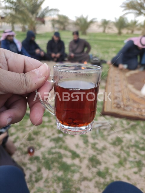 A close-up of a hand holding a cup of red tea, young Saudi Gulf Arabs having a fun popular Arab youth session in Saudi Arabia, a picnic in the open air, enjoying warm drinks, providing Gulf hospitality