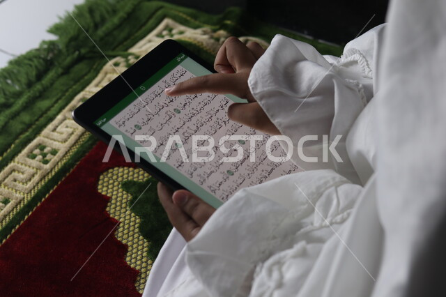 Worship and getting closer to God, reciting the Holy Qur’an through Islamic programs on the iPad, a close-up of a Saudi Arabian Gulf Muslim woman wearing a white prayer cover holding a tablet and pointing to the verses of the Qur’an, the electronic Quran 