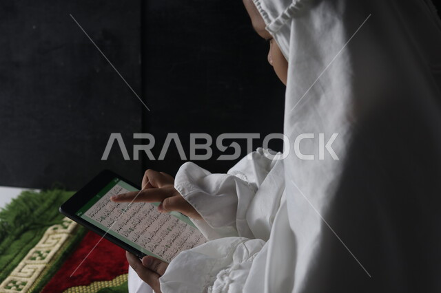 تلاوة القرآن الكريم عن طريق البرامج الاسلامية على الايباد, العبادة والتقرب إلى الله, صورة مقربة لإمرأة مسلمة عربية خليجية سعودية ترتدي غطاء الصلاة الأبيض تمسك جهاز التابليت وتقوم بالاشارة إلى آيات القرآن,المصحف الالكتروني المجود والمرتل, اسلاميات وعبادة