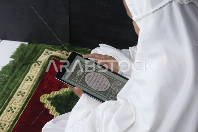 فضل وثواب قراءة وحفظ القرآن الكريم, صورة من الجانب لإمرأة عربية خليجية سعودية مسلمة ترتدي غطاء الصلاة الأبيض تقوم بقراءة القرآن عن طريق البرامج الإسلامية على التابليت, المصحف الالكتروني المجود والمرتل, اسلاميات وعبادة