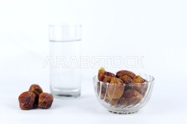 اتباع سنة رسول الله في كسر الصيام, صورة مقربة لصحن من التمور الجافة اللذيذة بجانب كأس الماء البارد, محاصيل زراعية وطنية محلية سعودية, وجبة غذائية ذات قيمة عالية في شهر رمضان, ضيافة رمضانية فاخرة, خلفية بيضاء