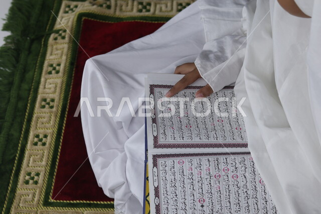 العبادة والتقرب إلى الله في شهر رمضان المبارك, مفهوم التربية على طريق و نور الإسلام، صورة مقربة لفتاة عربية خليجية سعودية صغيرة تجلس على سجادة الصلاة تحمل بيدها المصحف الشريف, تتبع كلام الله عز وجل ,تلاوة و تحفيظ القرآن الكريم