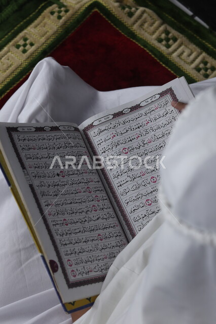 تلاوة و تحفيظ القرآن الكريم, مفهوم التربية على طريق و نور الإسلام، صورة مقربة لفتاة عربية خليجية سعودية صغيرة تجلس على سجادة الصلاة تحمل بيدها المصحف الشريف, تتبع كلام الله عز وجل ,العبادة والتقرب إلى الله في شهر رمضان المبارك