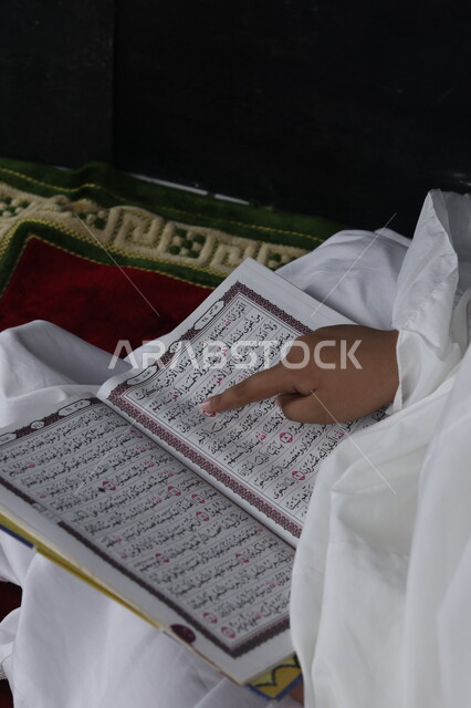 مفهوم التربية على طريق و نور الإسلام، صورة مقربة لفتاة عربية خليجية سعودية صغيرة تجلس على سجادة الصلاة تحمل بيدها المصحف الشريف, تتبع كلام الله عز وجل ,تلاوة و تحفيظ القرآن الكريم, العبادة والتقرب إلى الله في شهر رمضان المبارك