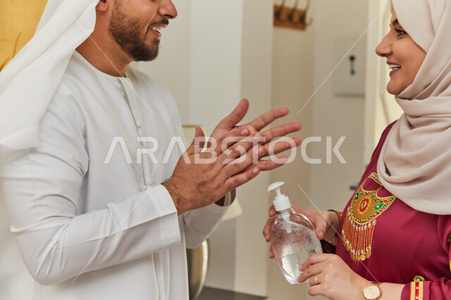 اتباع الإجراءات الإحترازيه للوقاية من الإصابه بفايروس كورونا، صورة مقربه لامرأة عربيه خليجية اماراتية ترتدي عباءه مطرزه تقوم بتعقيم يدي زوجها بعد عودته إلى المنزل، تجنب العدوى والحفاظ على السلامه من كوفيد-١٩.