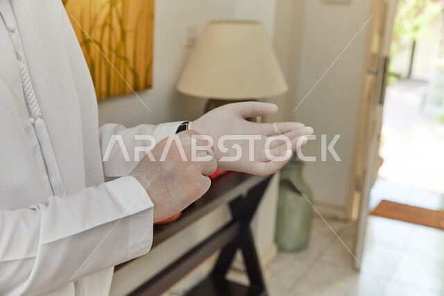 القيام بالتحضيرات والاستعداد للخروج من المنزل بزمن الكورونا، صورة مقربه ليدين رجل عربي خليجي اماراتي يقوم بإرتداء القفازات الطبيه المعقمة للحمايه من كوفيد-١٩، الحفاظ على السلامة وتجنب العدوى بالفايروسات.