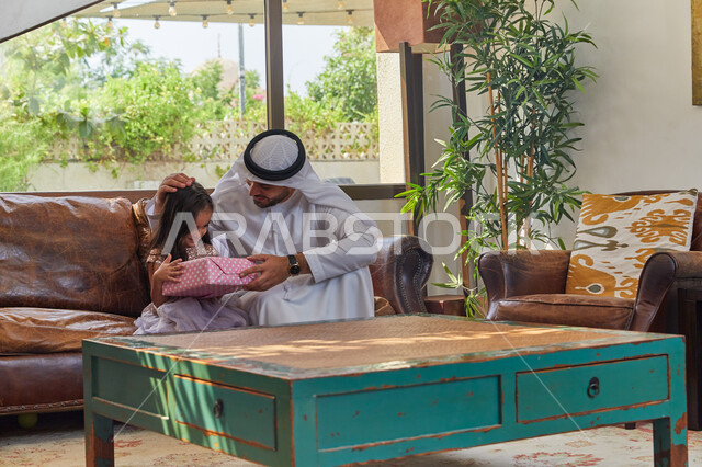  الأعياد والمناسبات الإسلامية في المجتمع الاماراتي، أجواء عائلية مليئة بالحب، ايماءات الفرح بتقديم الهدايا، رجل عربي اماراتي خليجي يرتدي الكندورة و الغترة الاماراتية يجلس في غرفة المعيشة مع ابنته، الضحك والتسامر مع الأبناء، الاحتفال بقدوم العيد السعيد