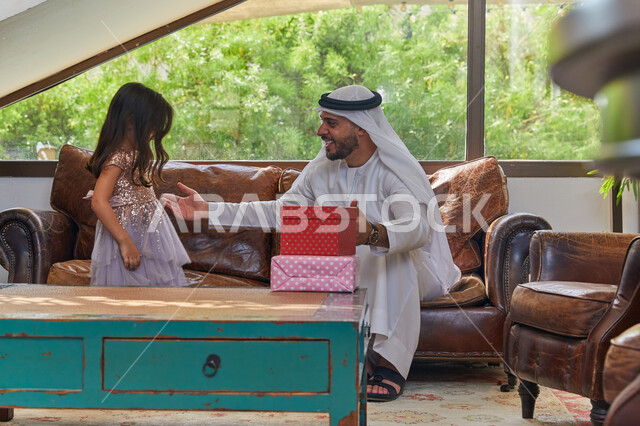 التهنئة بقدوم العيد السعيد، تقديم الهدايا في الأعياد والمناسبات الإسلامية، صورة مقربه لرجل عربي اماراتي خليجي يرتدي الكندورة و الغترة الاماراتية يجلس في غرفة المعيشة مع ابنته، مفهوم التربية بالحب والحنان، قضاء أوقات ممتعه مع الابنه بإيماءات السعادة