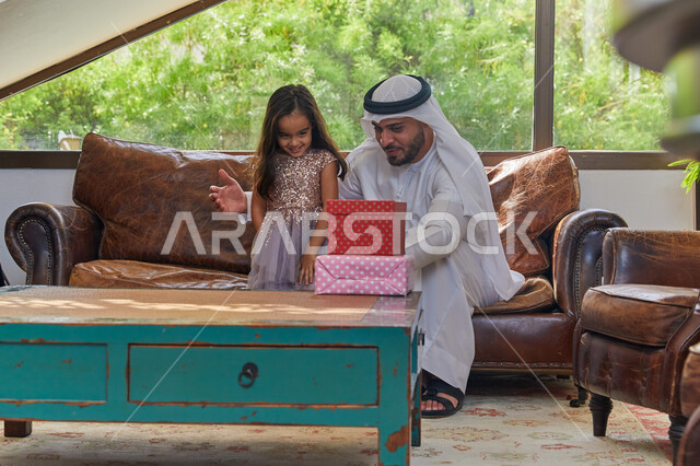 ايماءات الفرح  بتقديم الهدايا في الأعياد والمناسبات الإسلامية، صورة مقربه لرجل عربي اماراتي خليجي يرتدي الكندورة و الغترة الاماراتية يجلس في غرفة المعيشة مع ابنته، الضحك والتسامر مع الأبناء، الاحتفال بقدوم العيد السعيد، مفهوم التربية بالحب والحنان