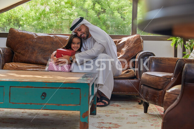 The concept of education with love and tenderness, presenting gifts on Islamic holidays and occasions, a close-up image of an Arab Emirati Gulf man wearing an Emirati kandura and ghutra sitting in the living room featuring his daughter, laughing and commu