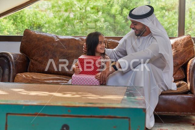تقديم الهدايا في الأعياد والمناسبات الإسلامية، صورة مقربه لرجل عربي اماراتي خليجي يرتدي الكندورة و الغترة الاماراتية يجلس في غرفة المعيشة مع ابنته، أجواء عائلية مليئة بالحب، الضحك والتسامر مع الأبناء بإيماءات الفرح، الاحتفال بقدوم العيد السعيد