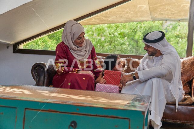 الضحك والتسامر مع الأبناء، أجواء عائلية سعيدة مليئة بالحب, الاحتفال بقدوم العيد المبارك، تقديم علب الهدايا للإبنه ، عائلة عربية خليجية سعودية يجلسون في غرفة المعيشة بإيماءات السعادة والفرح، ارتداء الزي الإماراتي في الأعياد والمناسبات الإسلامية
