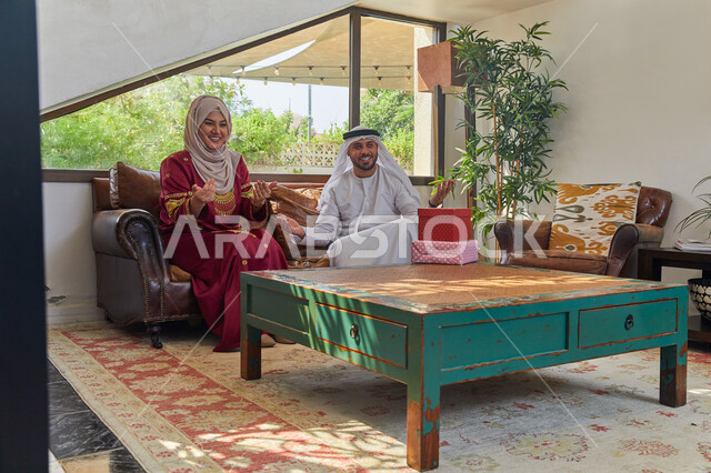الاحتفال بقدوم العيد المبارك، جلسات مميزة مع الأسرة، تقديم الهدايا في الأعياد والمناسبات الاسلامية، زوجان عربيان اماراتيان خليجيان يجلسان في غرفة المعيشة بإيماءات السعادة والفرح، أجواء عائلية سعيدة مليئة بالحب