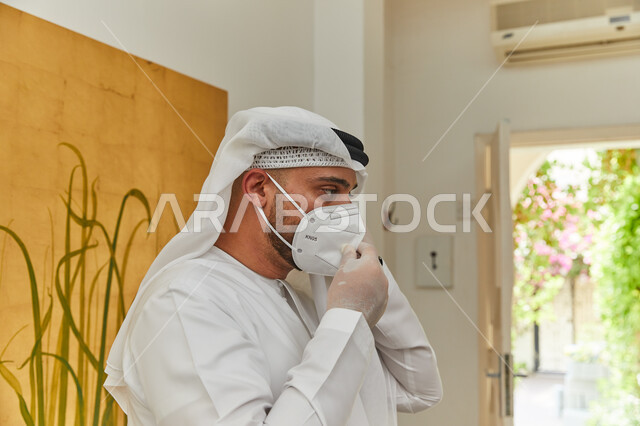 الحفاظ على السلامة وتجنب العدوى بالفايروسات، القيام بالتحضيرات والاستعداد للخروج من المنزل بزمن الكورونا، صورة مقربه لرجل عربي خليجي اماراتي يقوم بوضع الكمامة ويرتدي الزي التقليدي والقفازات الطبيه المعقمة للحمايه من كوفيد-١٩.