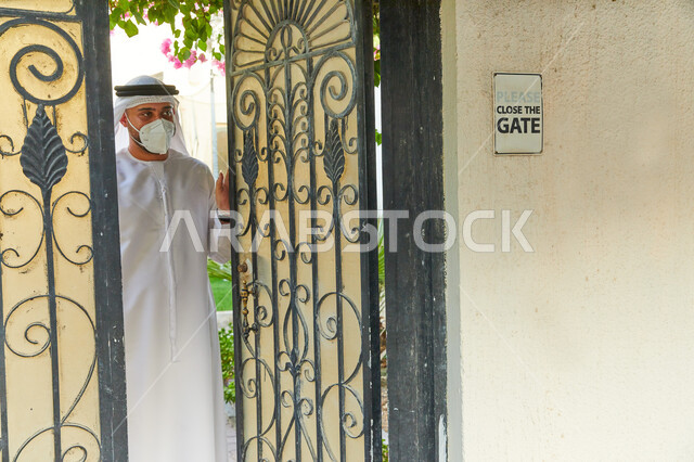 فلل و شقق سكنية فاخرة في الامارات، رجل عربي خليجي اماراتي يرتدي الكندورة و الغترة يقوم بفتح الباب واستقبال احد ما، انتظار مندوب الطلبيات والمشتريات الإلكترونيه الاونلاين، الالتزام بالاجراءات الاحترازية و ارتداء الكمامة للوقاية من الامراض وفايروس كورونا.