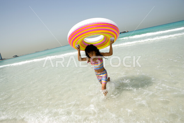 قضاء أوقات ممتعة في البحر, منتجعات شاطئية في الإمارات, فتاة عربية إماراتية خليجية تجري على الشاطئ وتحمل بيدها عوامة السباحة, أنشطة ترفيهية مسلية في العطلة, الاستمتاع بالفعاليات الصيفية