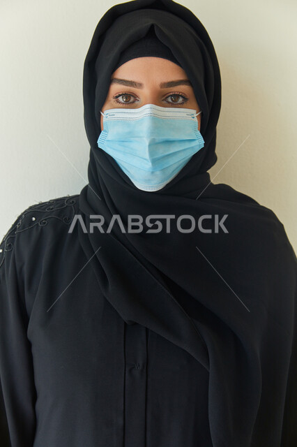 ايماءات تدل على النظر إلى الكاميرا، بورتريه مقرب لشابه عربية خليجية اماراتية محجبة ترتدي عباءه سوداء وتضع الكمامة لتجنب تفشي فايروس كورونا، إتباع الإجراءات الوقايه والسلامة للحمايه من العدوى بكوفيد-١٩، خلفية بيضاء.