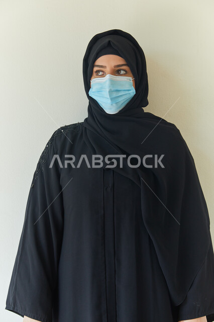 ايماءات تدل على النظر إلى شيء ما، بورتريه مقرب لشابه عربية خليجية اماراتية محجبة ترتدي عباءه سوداء وتضع الكمامة لتجنب تفشي فايروس كورونا، إتباع الإجراءات الوقايه والسلامة للحمايه من العدوى بكوفيد-١٩، خلفية بيضاء.